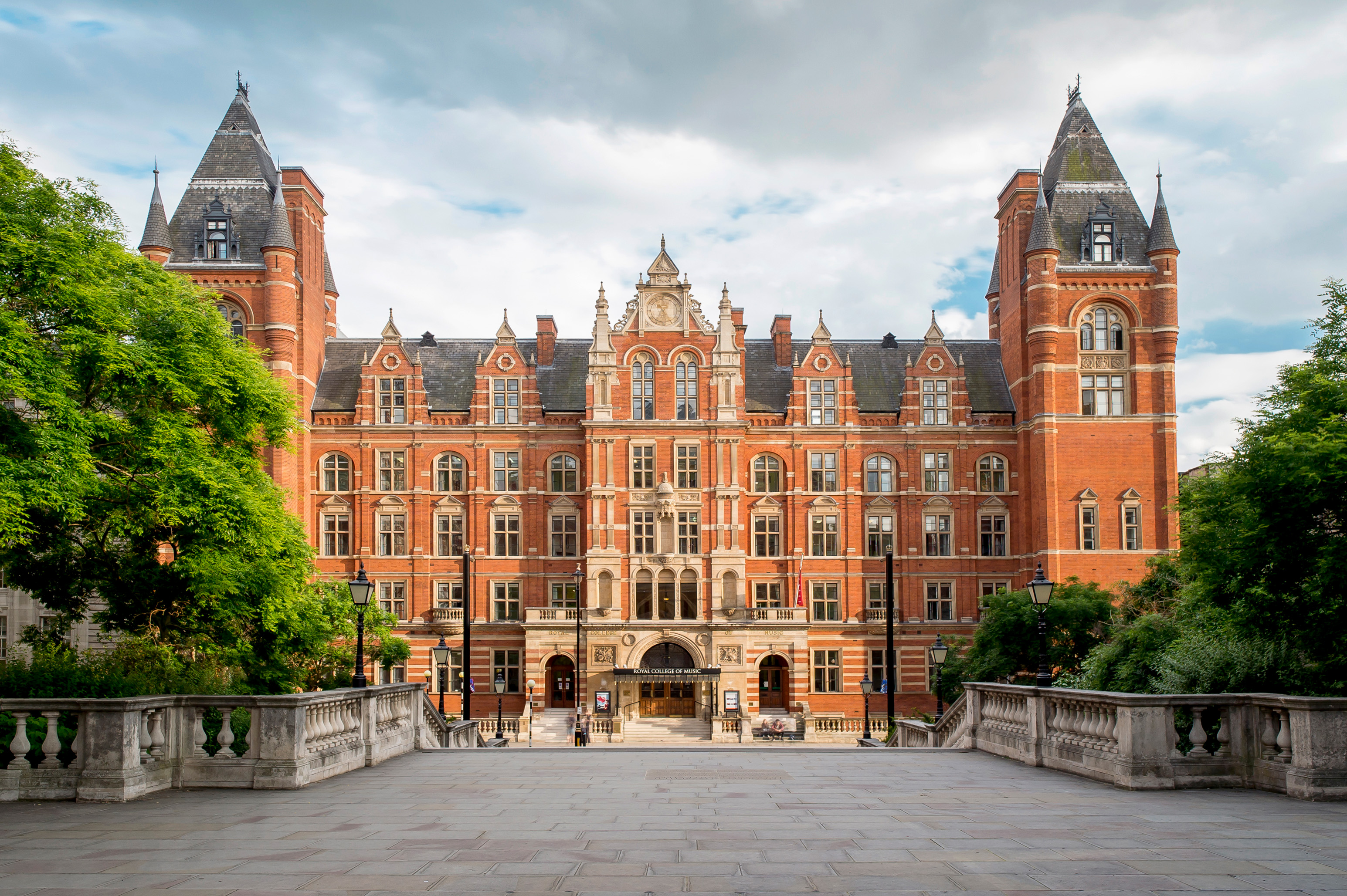 A red brick building, with two towers, with steps leading up to the entrance, framed by a green trees on both sides with a cloudy sky.